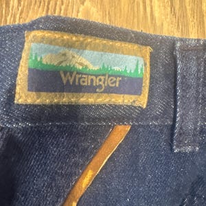 Puede incluir: Primer plano de una tela vaquera azul oscuro con un parche de cuero marr&oacute;n. El parche tiene un fondo blanco con una escena de monta&ntilde;a verde y la palabra "Wrangler" en blanco.