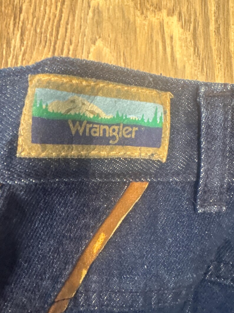 Puede incluir: Primer plano de una tela vaquera azul oscuro con un parche de cuero marr&oacute;n. El parche tiene un fondo blanco con una escena de monta&ntilde;a verde y la palabra "Wrangler" en blanco.