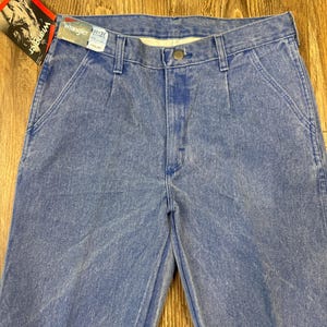 May include: Light blue Wrangler jeans with a tag attached. The jeans are a classic style with a button closure and pockets. The tag shows a person wearing a cowboy hat.
