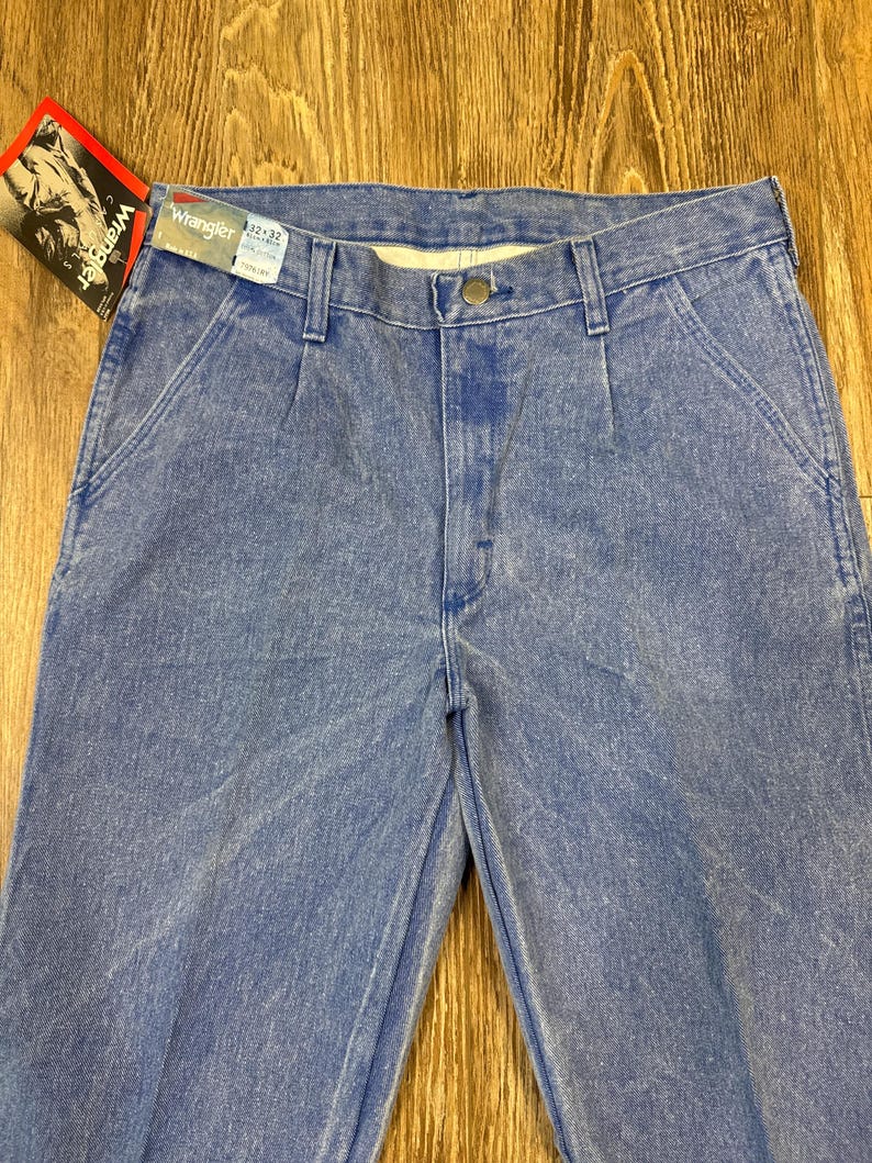 May include: Light blue Wrangler jeans with a tag attached. The jeans are a classic style with a button closure and pockets. The tag shows a person wearing a cowboy hat.