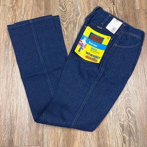 K&ouml;nnte beinhalten: Dunkelblaue Wrangler Bootcut-Jeans mit gelb-braunem Etikett. Das Etikett tr&auml;gt die Aufschrift "Boot Jean" und "Wrangler" mit einer Grafik einer Person. Die Jeans liegen auf einer Holzoberfl&auml;che.