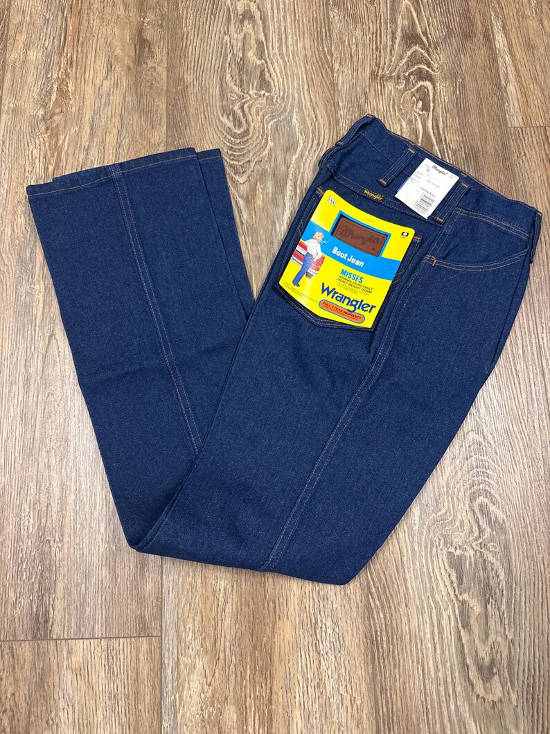 K&ouml;nnte beinhalten: Dunkelblaue Wrangler Bootcut-Jeans mit gelb-braunem Etikett. Das Etikett tr&auml;gt die Aufschrift "Boot Jean" und "Wrangler" mit einer Grafik einer Person. Die Jeans liegen auf einer Holzoberfl&auml;che.