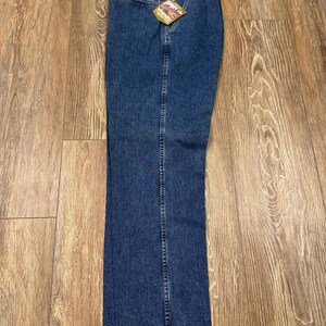 May include: Blue denim jeans with a straight leg design. The jeans have a classic five-pocket style, a zipper fly, and a button closure. A tag is attached to the jeans. The jeans are laid flat on a wooden surface.