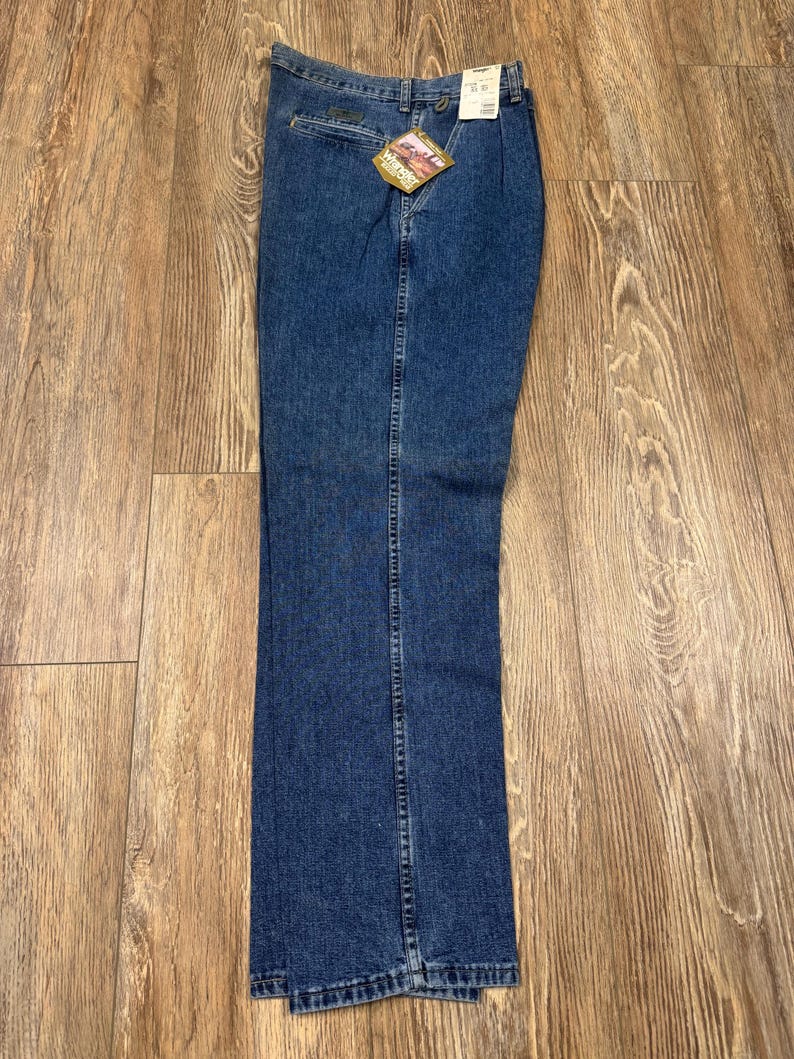 May include: Blue denim jeans with a straight leg design. The jeans have a classic five-pocket style, a zipper fly, and a button closure. A tag is attached to the jeans. The jeans are laid flat on a wooden surface.
