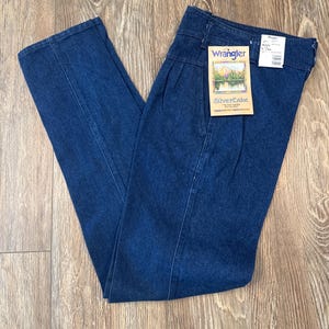 Puede incluir: Vaqueros Wrangler de mezclilla azul oscuro con cintura alta y pliegues. Los vaqueros tienen un dise&ntilde;o de pernera recta y una etiqueta que dice "Wrangler SilverLake". Los vaqueros est&aacute;n doblados sobre una superficie de madera.