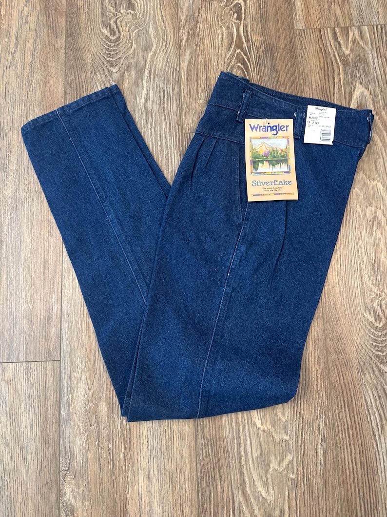Puede incluir: Vaqueros Wrangler de mezclilla azul oscuro con cintura alta y pliegues. Los vaqueros tienen un dise&ntilde;o de pernera recta y una etiqueta que dice "Wrangler SilverLake". Los vaqueros est&aacute;n doblados sobre una superficie de madera.
