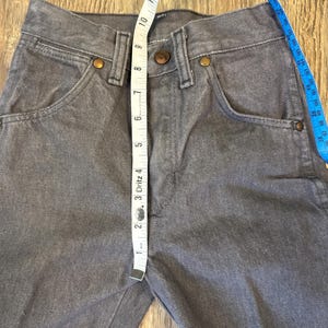 May include: Close-up of gray denim jeans with a tape measure showing a measurement. The jeans have classic five-pocket styling with copper-colored rivets and a button closure. The tape measure shows a measurement of approximately 1 inch.