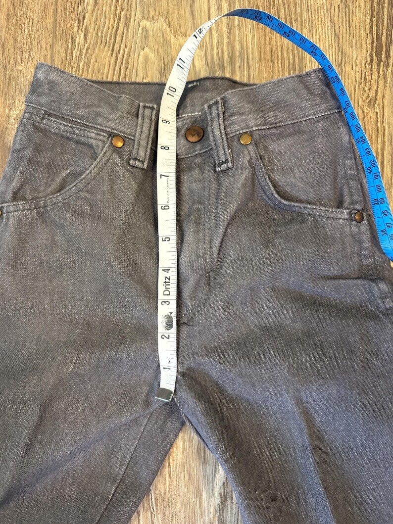 May include: Close-up of gray denim jeans with a tape measure showing a measurement. The jeans have classic five-pocket styling with copper-colored rivets and a button closure. The tape measure shows a measurement of approximately 1 inch.