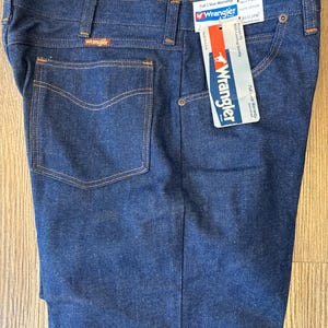 May include: A pair of dark blue denim jeans with a brown leather Wrangler label on the back pocket. The jeans have a tag attached with the text "Wrangler" and "Full 'N' Wear Warranty". The tag also indicates the size is 34 x 32, 100% cotton, and the style number is 9331PW.
