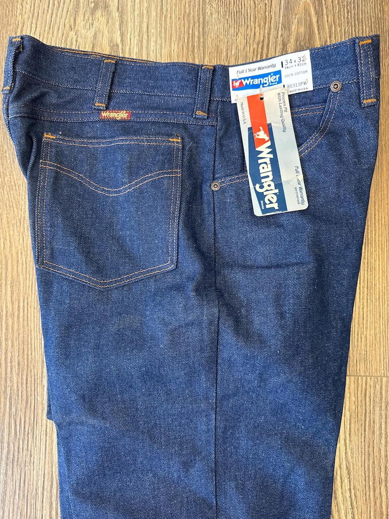 May include: A pair of dark blue denim jeans with a brown leather Wrangler label on the back pocket. The jeans have a tag attached with the text "Wrangler" and "Full 'N' Wear Warranty". The tag also indicates the size is 34 x 32, 100% cotton, and the style number is 9331PW.