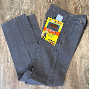 May include: Gray Wrangler Cowboy Cut jeans with a yellow and orange tag. The tag reads "prorodeo" and "Cowboy Cut" with a graphic of a cowboy. The jeans are folded on a wooden surface.