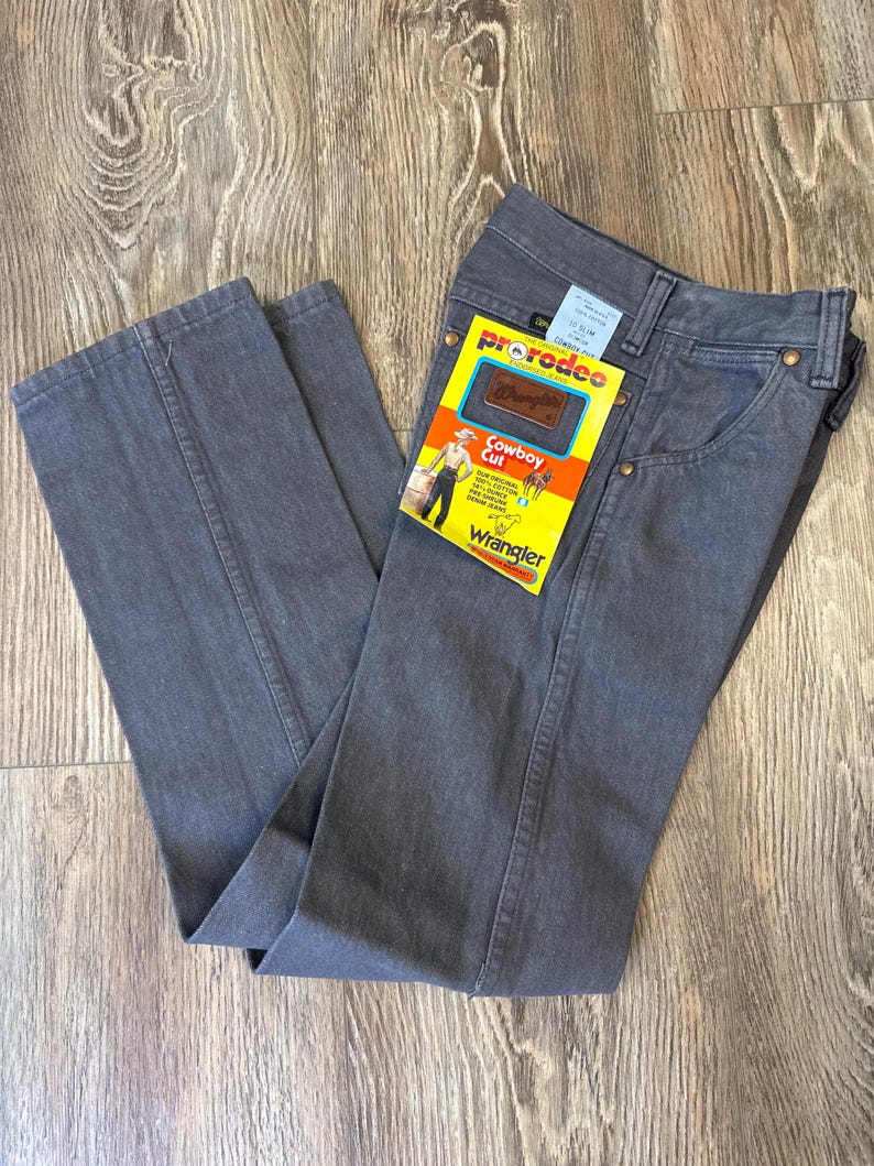 May include: Gray Wrangler Cowboy Cut jeans with a yellow and orange tag. The tag reads "prorodeo" and "Cowboy Cut" with a graphic of a cowboy. The jeans are folded on a wooden surface.