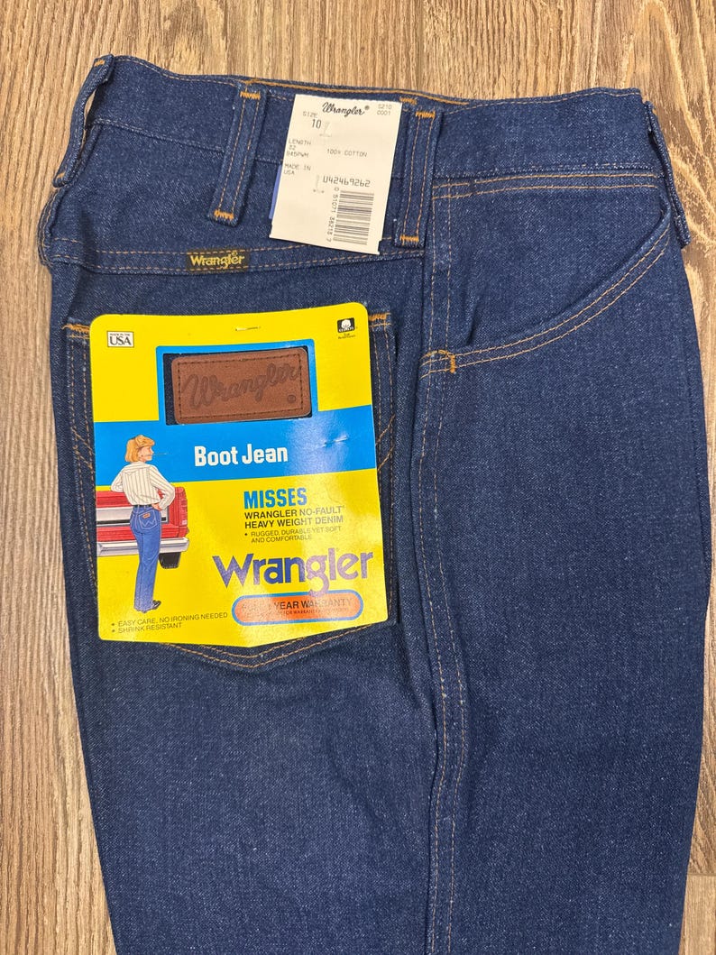 K&ouml;nnte beinhalten: Dunkelblaue Wrangler Bootcut-Jeans mit gelb-braunem Etikett. Das Etikett lautet "Boot Jean" und "Misses Wrangler No-Fault Heavy Weight Denim". Die Jeans haben ein klassisches F&uuml;nf-Taschen-Design und ein Wrangler-Label.