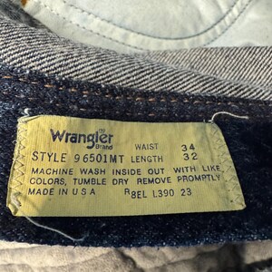 May include: Close-up of a Wrangler brand jeans tag. The yellow tag displays black text with the style, waist size 86 cm, and length 81 cm. Washing instructions and "Made in USA" are also printed on the tag.