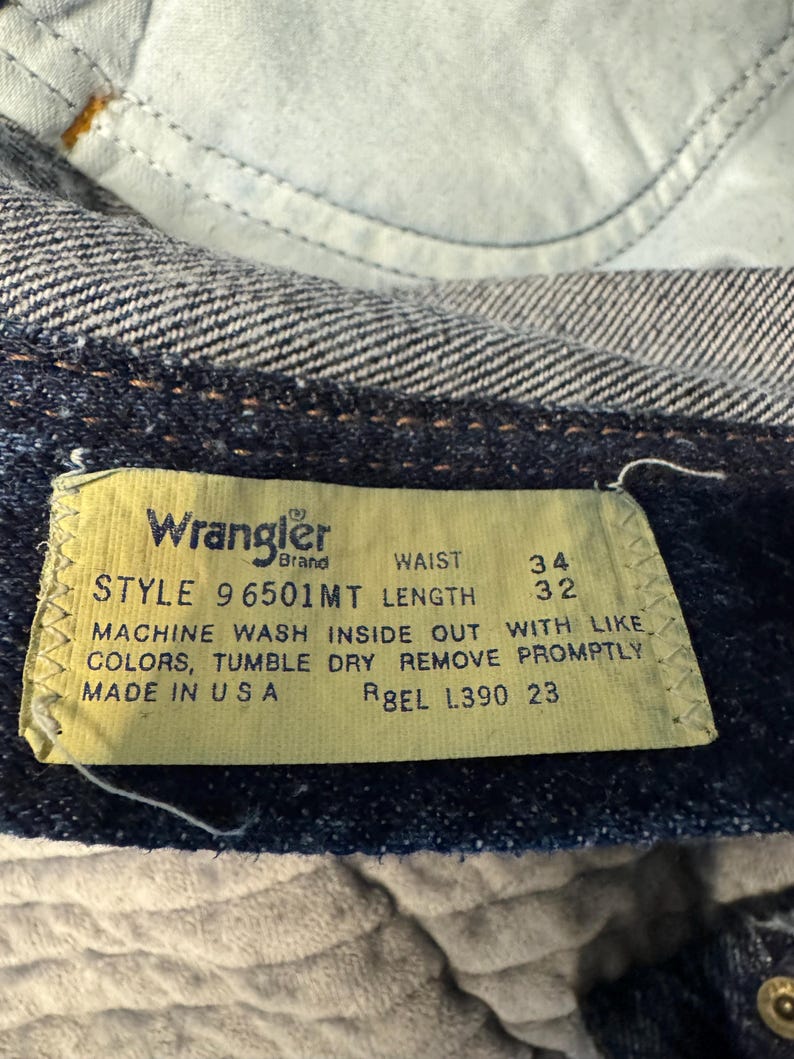May include: Close-up of a Wrangler brand jeans tag. The yellow tag displays black text with the style, waist size 86 cm, and length 81 cm. Washing instructions and "Made in USA" are also printed on the tag.