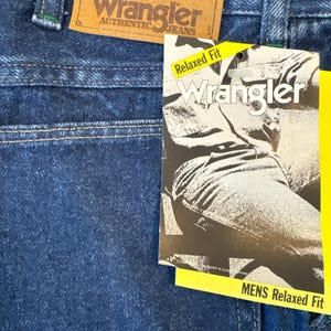 May include: Close-up of blue denim Wrangler jeans. A brown leather tag displays the Wrangler logo and the words "AUTHENTIC JEANS." A yellow tag reads "Relaxed Fit" and "MENS Relaxed Fit."