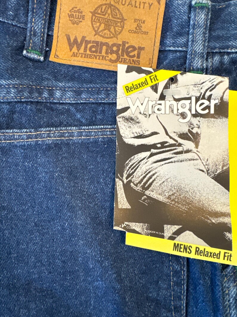 May include: Close-up of blue denim Wrangler jeans. A brown leather tag displays the Wrangler logo and the words "AUTHENTIC JEANS." A yellow tag reads "Relaxed Fit" and "MENS Relaxed Fit."