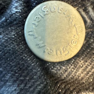 Puede incluir: Un bot&oacute;n de metal plateado con el texto "Wrangler" y "Genuine" alrededor del borde.