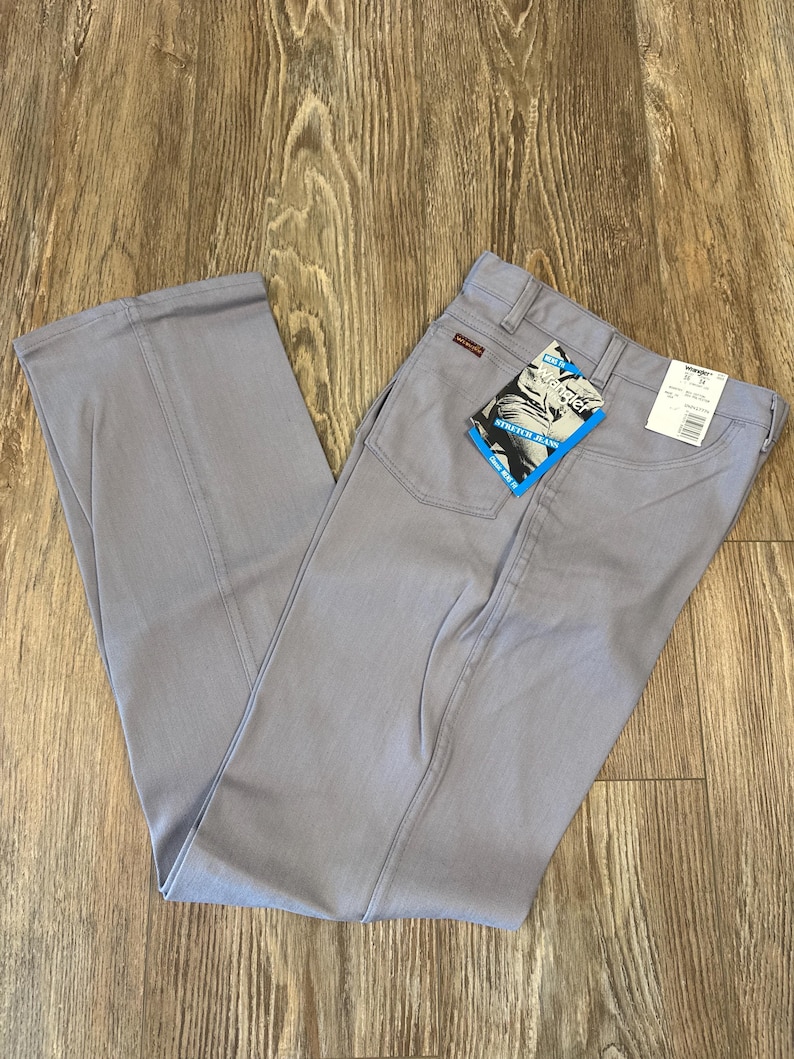 May include: Light gray Wrangler jeans with a straight leg design. The jeans have a classic five-pocket style, a zipper fly, and a Wrangler label on the back pocket. A blue and white tag is attached to the jeans.