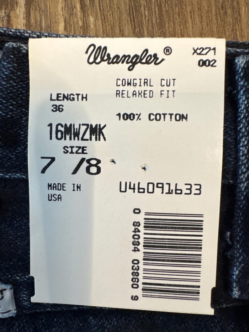 May include: A white tag with the Wrangler logo and text. The tag reads "COWGIRL CUT RELAXED FIT" and "100% COTTON". It also indicates a length of 36 inches and a size of 7/8. The tag is marked "MADE IN USA".