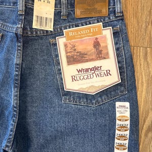 May include: A pair of blue denim jeans with a brown leather patch that says "Wrangler Rugged Wear". The jeans are size 34x32 and have a relaxed fit.