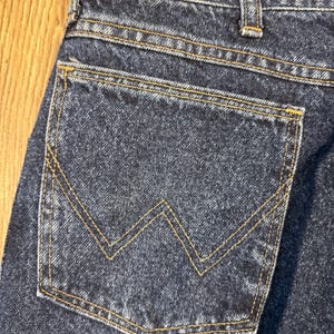 Puede incluir: Primer plano del bolsillo trasero de unos vaqueros de mezclilla azul oscuro. El bolsillo presenta una costura distintiva en forma de "W" en hilo amarillo-dorado. Los vaqueros est&aacute;n hechos de un tejido de mezclilla resistente.
