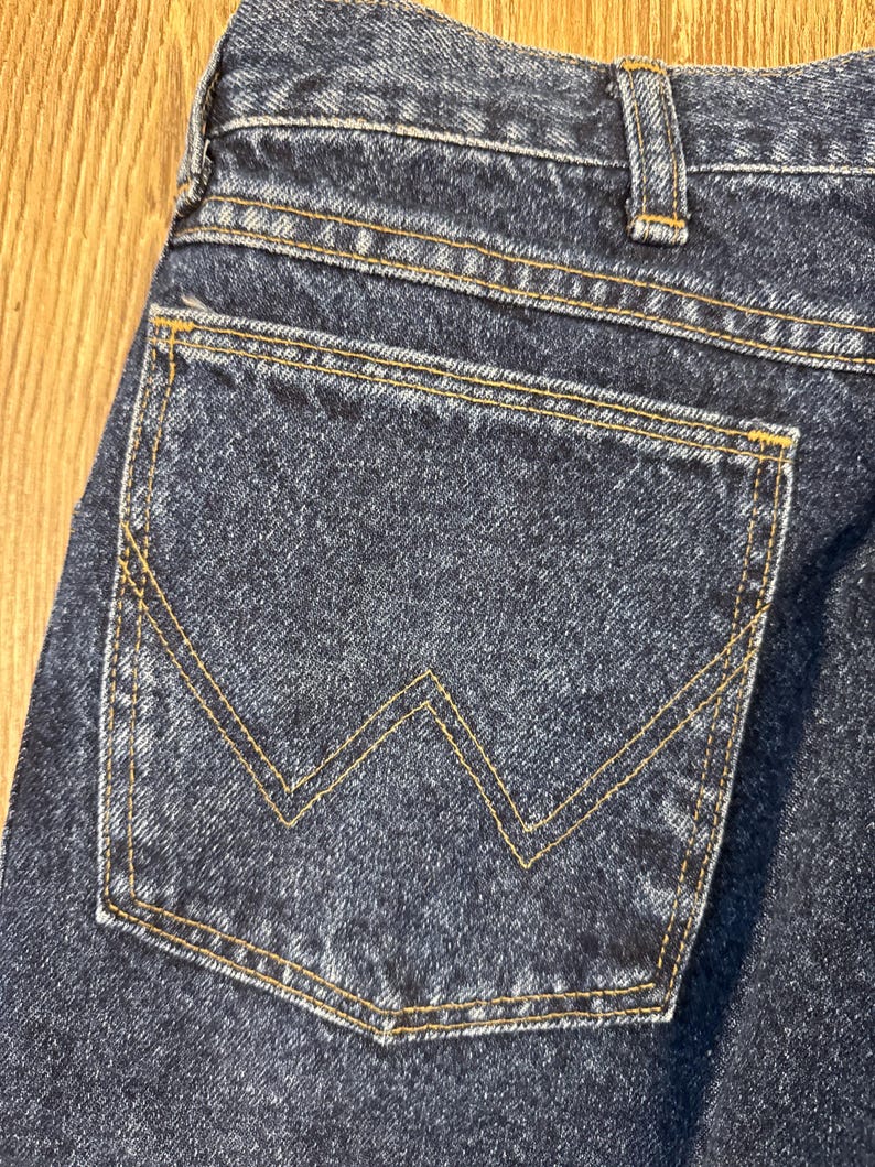 Puede incluir: Primer plano del bolsillo trasero de unos vaqueros de mezclilla azul oscuro. El bolsillo presenta una costura distintiva en forma de "W" en hilo amarillo-dorado. Los vaqueros est&aacute;n hechos de un tejido de mezclilla resistente.
