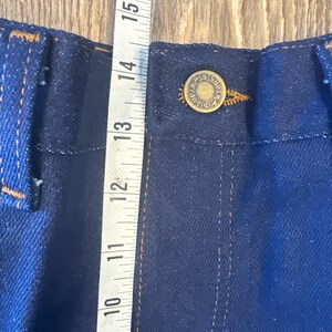 K&ouml;nnte beinhalten: Nahaufnahme eines dunkelblauen Jeansstoffes mit einem kupfernen Knopf und N&auml;hten. Links im Bild ist ein Ma&szlig;band zu sehen, das Ma&szlig;e in Zoll anzeigt.