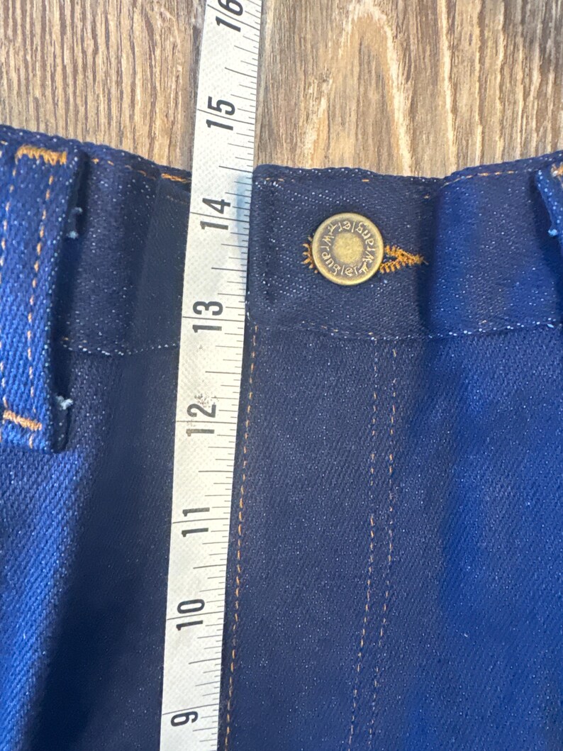 K&ouml;nnte beinhalten: Nahaufnahme eines dunkelblauen Jeansstoffes mit einem kupfernen Knopf und N&auml;hten. Links im Bild ist ein Ma&szlig;band zu sehen, das Ma&szlig;e in Zoll anzeigt.