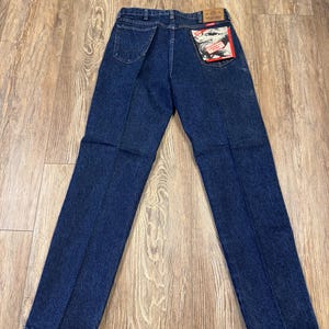 May include: Dark blue denim jeans with a classic five-pocket design. The jeans have a straight leg cut and a branded patch on the back pocket. The jeans are laid flat on a wooden surface.