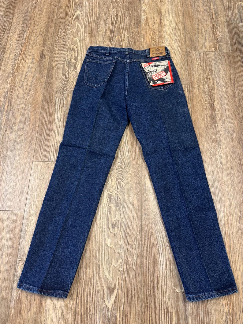 May include: Dark blue denim jeans with a classic five-pocket design. The jeans have a straight leg cut and a branded patch on the back pocket. The jeans are laid flat on a wooden surface.