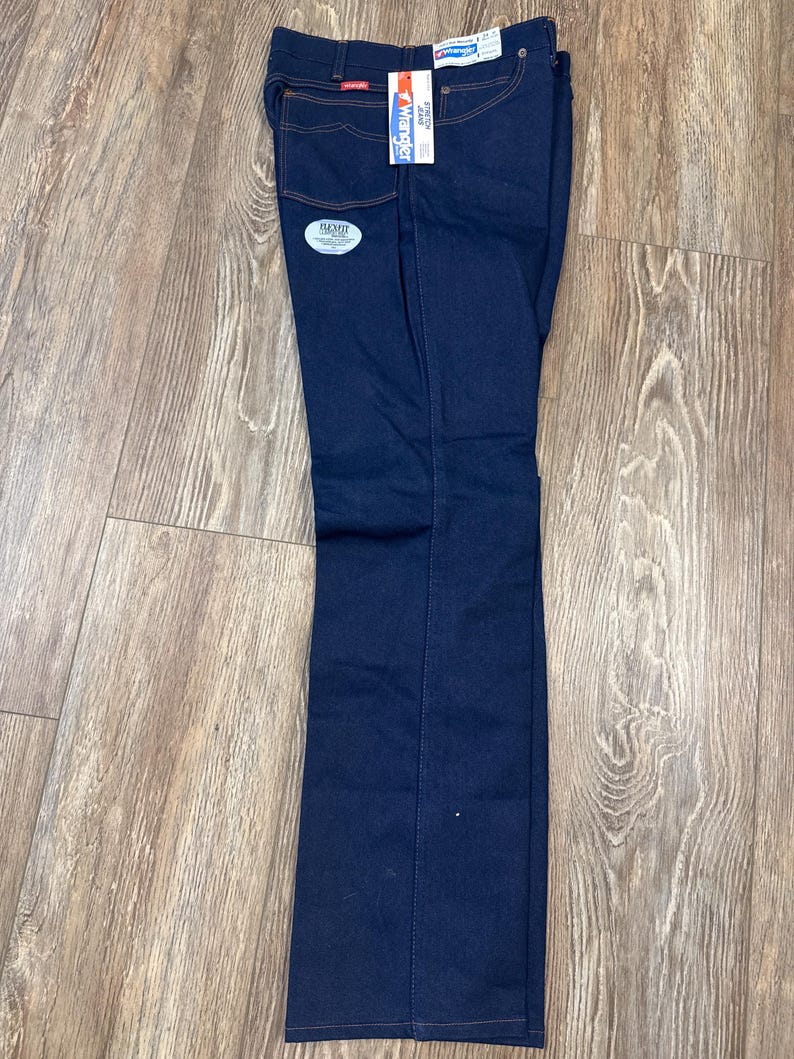 May include: Dark blue Wrangler jeans with orange stitching and a Flex Fit tag. The jeans have a straight leg cut and are folded neatly. A white tag with the Wrangler logo is attached to the back pocket.