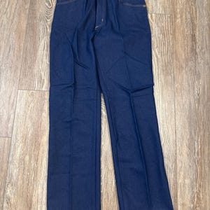 May include: Dark blue denim jeans with a straight leg design. The jeans have a classic five-pocket style, a button and zipper closure, and a Wrangler tag attached. The stitching is a contrasting tan color.