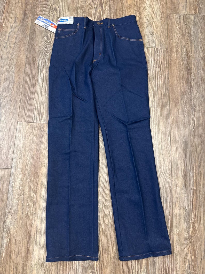 May include: Dark blue denim jeans with a straight leg design. The jeans have a classic five-pocket style, a button and zipper closure, and a Wrangler tag attached. The stitching is a contrasting tan color.