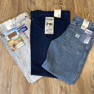 May include: Three pairs of Wrangler jeans in various washes: light gray "Hurricane Wash", dark blue, and light blue "Wrangler Blues". Each pair has tags attached. The jeans are laid out on a wooden surface.