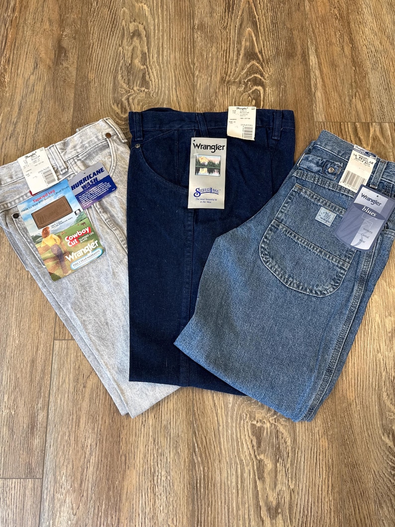 May include: Three pairs of Wrangler jeans in various washes: light gray "Hurricane Wash", dark blue, and light blue "Wrangler Blues". Each pair has tags attached. The jeans are laid out on a wooden surface.
