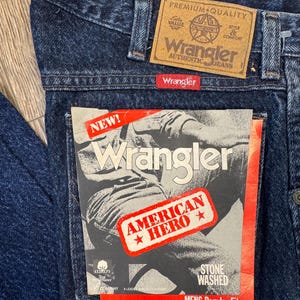 May include: Close-up of dark blue Wrangler jeans with a brown leather patch and a red Wrangler tag. The jeans have a "NEW!" tag with "AMERICAN HERO" and "STONE WASHED" text. The jeans are "MENS Regular Fit".