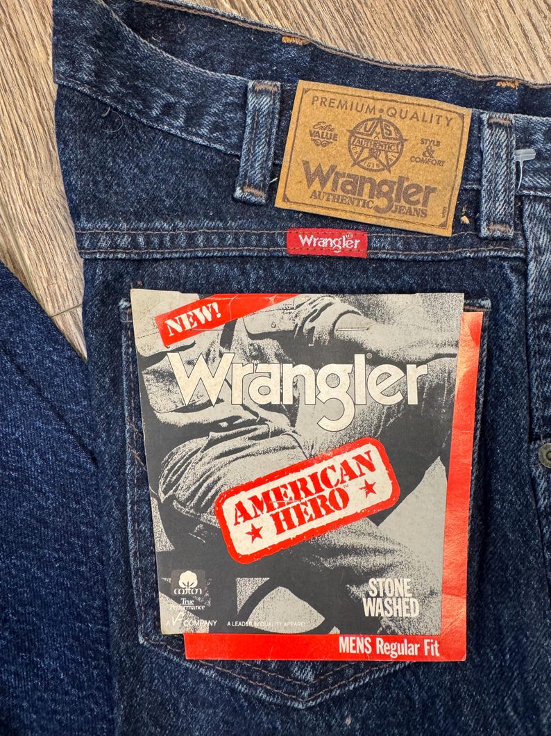 May include: Close-up of dark blue Wrangler jeans with a brown leather patch and a red Wrangler tag. The jeans have a "NEW!" tag with "AMERICAN HERO" and "STONE WASHED" text. The jeans are "MENS Regular Fit".