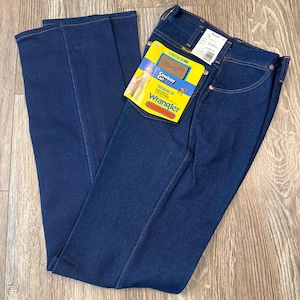 May include: Dark blue Wrangler Cowboy Cut jeans with a regular fit. The jeans have a yellow and blue tag attached with the Wrangler logo and product information. The jeans are made of stretch denim.