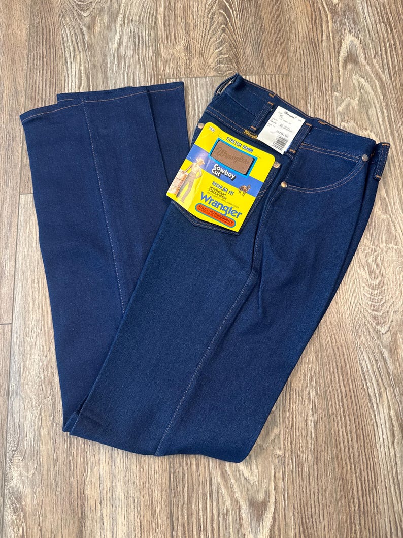 May include: Dark blue Wrangler Cowboy Cut jeans with a regular fit. The jeans have a yellow and blue tag attached with the Wrangler logo and product information. The jeans are made of stretch denim.