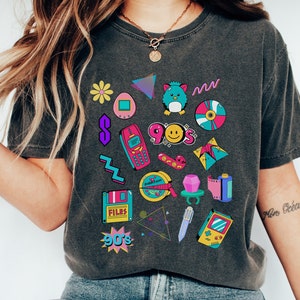 May include: A dark gray t-shirt with a 90s theme featuring a variety of colorful retro icons, including a Tamagotchi, a Game Boy, a floppy disk, a cassette tape, a cell phone, and a smiley face.
