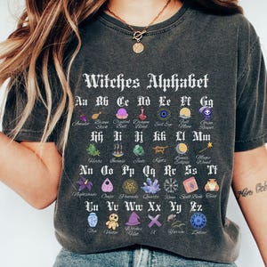 Pode incluir: Uma camiseta cinza escura com um gráfico branco de um alfabeto de bruxas. O alfabeto é ilustrado com vários símbolos e ícones de bruxas. O texto diz "Witches Alphabet".