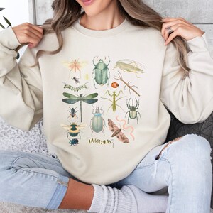 May include: A beige sweatshirt featuring a colourful insect print. The print includes a variety of insects, such as butterflies, beetles and dragonflies.
