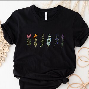 Może przedstawiać: Czarny T-shirt z rzędem haftowanych kwiatów w różnych kolorach, w tym różowym, pomarańczowym, żółtym, niebieskim i fioletowym.