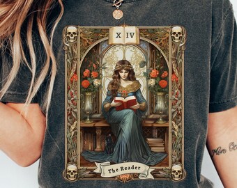 May include: Dark grey t-shirt featuring a tarot card design. The graphic depicts a woman reading a book, framed by ornate details, skulls, and the text "The Reader." The card is labeled "XIV."