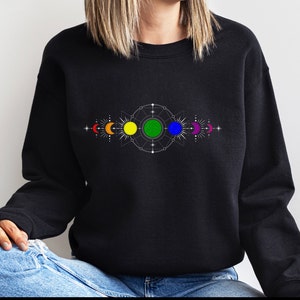 Pode incluir: Camisola de cor preta com um design de lua e sol arco-íris. O design apresenta um padrão geométrico com um círculo verde no centro.