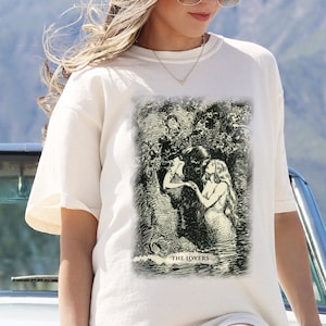 Pode incluir: Camiseta branca com uma ilustra&ccedil;&atilde;o em preto e branco de uma carta de tar&ocirc; que representa Os Amantes. O cart&atilde;o mostra duas figuras se abra&ccedil;ando em um ambiente de floresta. O texto "THE LOVERS" &eacute; impresso abaixo da ilustra&ccedil;&atilde;o.