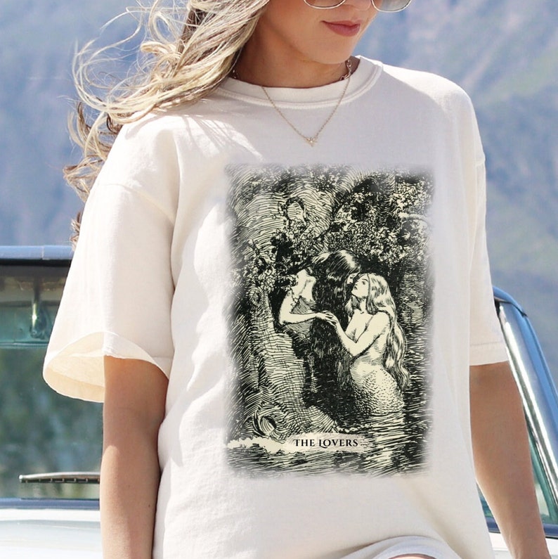 Pode incluir: Camiseta branca com uma ilustra&ccedil;&atilde;o em preto e branco de uma carta de tar&ocirc; que representa Os Amantes. O cart&atilde;o mostra duas figuras se abra&ccedil;ando em um ambiente de floresta. O texto "THE LOVERS" &eacute; impresso abaixo da ilustra&ccedil;&atilde;o.