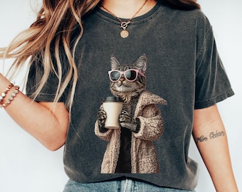 May include: Dark grey t-shirt with a cat illustration wearing pink sunglasses and a coat, holding a coffee cup. The cat has a relaxed, stylish appearance. This is a casual wear item.
