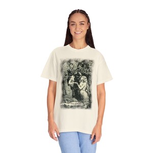 Pode incluir: Uma camiseta branca com uma ilustra&ccedil;&atilde;o vintage de um casal se abra&ccedil;ando em um cen&aacute;rio de floresta. A ilustra&ccedil;&atilde;o &eacute; em tons de verde e preto e inclui o texto "THE LOVERS".